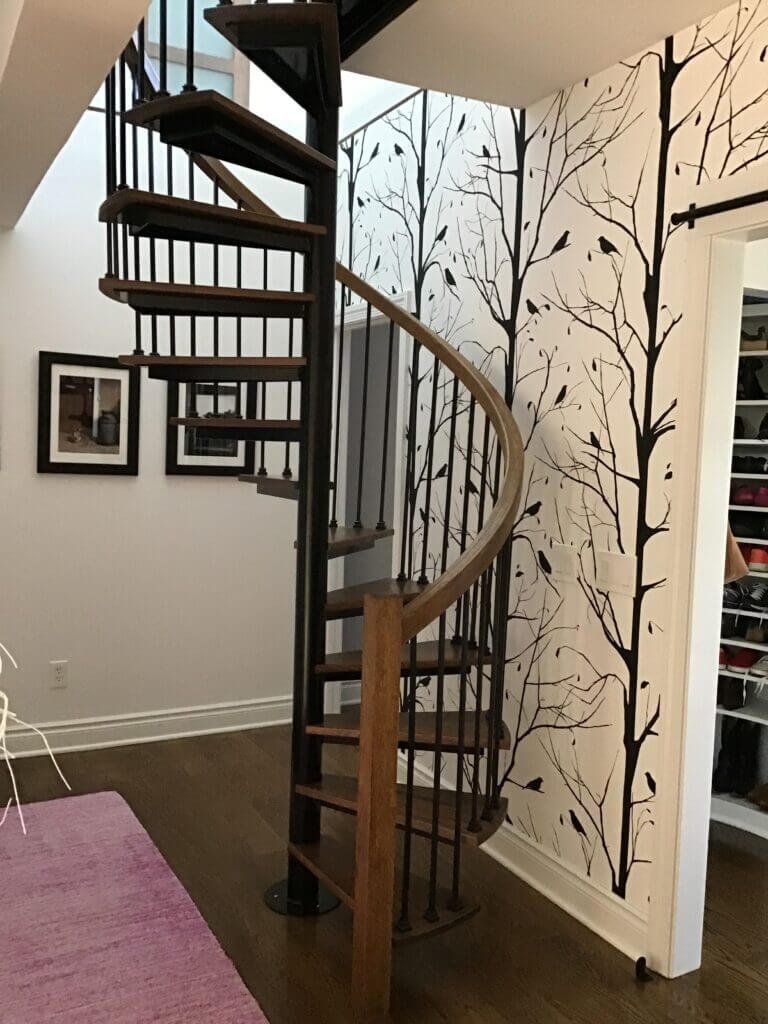 Spiral staircase with wood and black iron parts, in front of a wall with a black tree mural