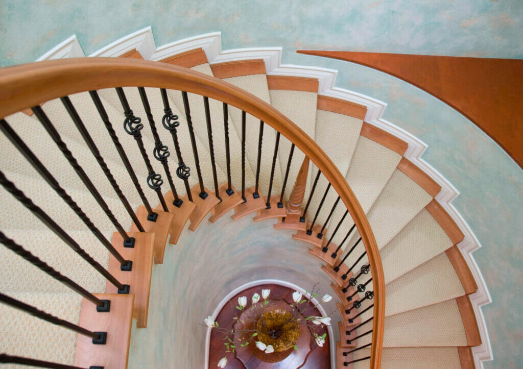 looking down on a spiral flight of stairs with a light wood banister and wood treads with a carpet runner